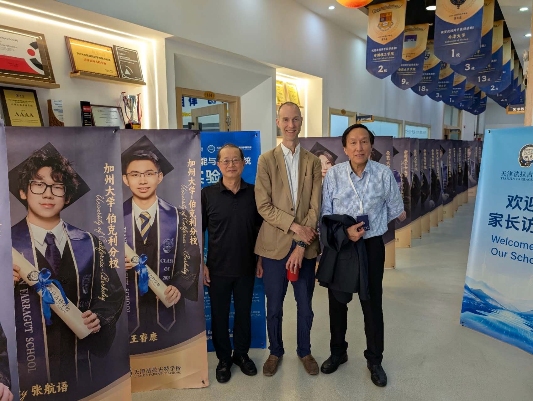 Chuck Eesley with two CFCC organizers in a hallway at Tianjin Farragut School, with student graduation banners showing US and UK university placements behind them