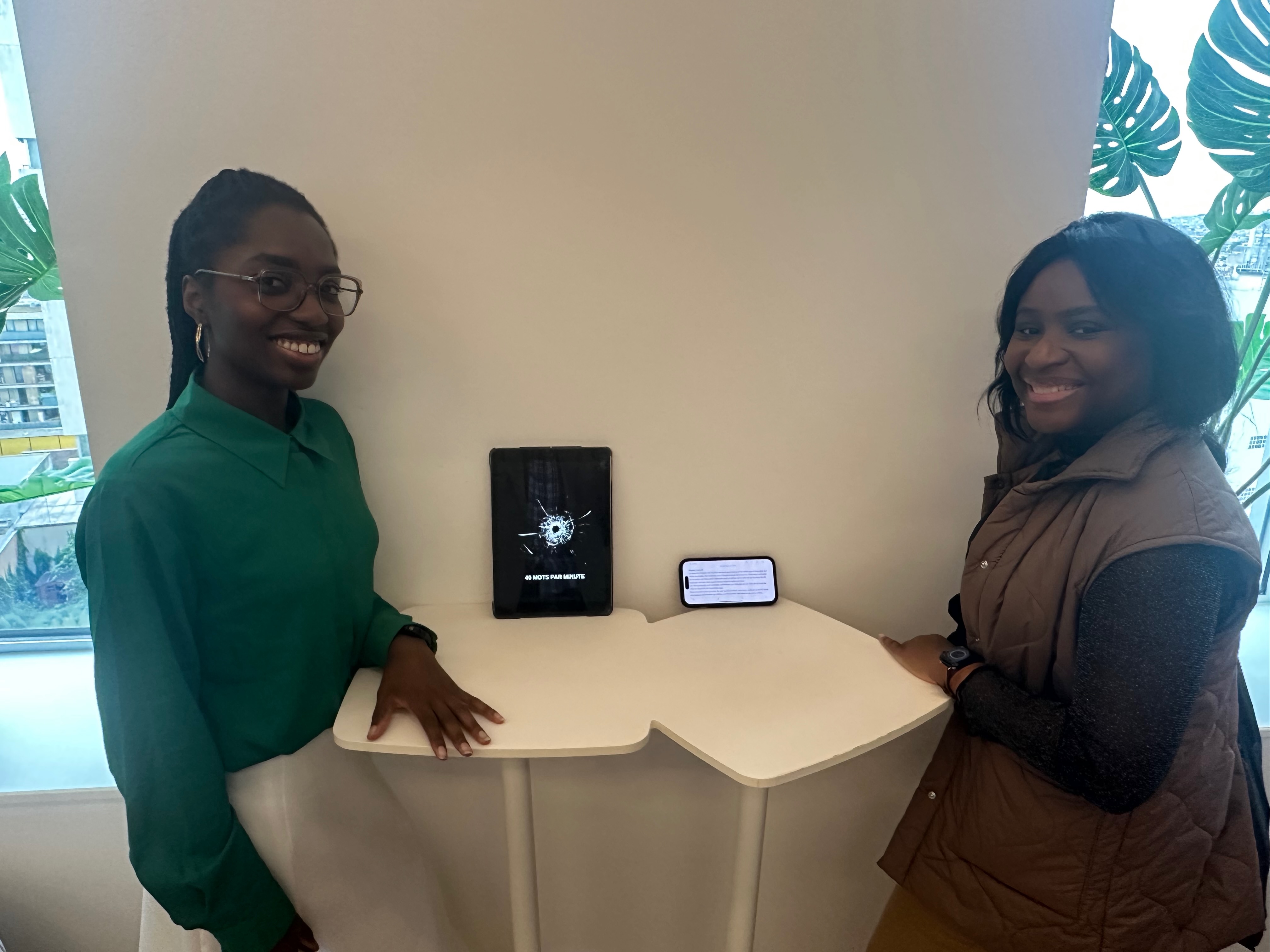 Two L'Ascenseur participants engaging with one of the fifteen Improbable Chair exhibition installations — an iPad showing a bullet-hole graphic and the text '40 mots par minute'