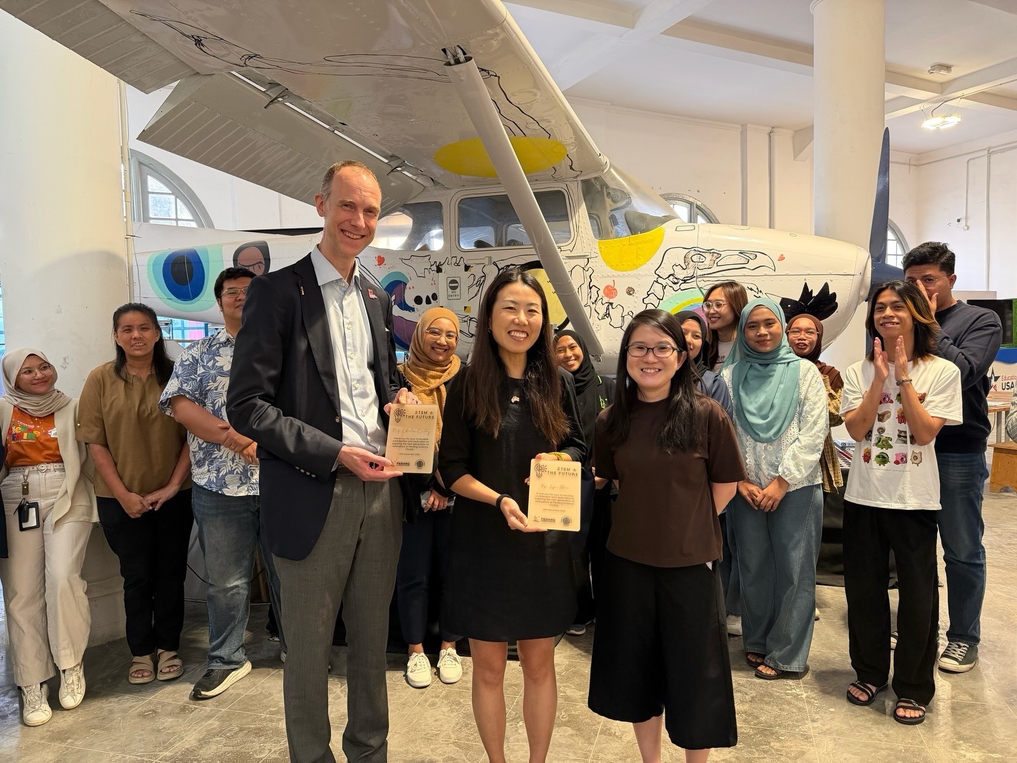 Chuck Eesley and Lijie Zhou holding appreciation plaques surrounded by Penang Science Cluster students and staff in the American Tech Corner, with a student-painted Cessna aircraft in the background