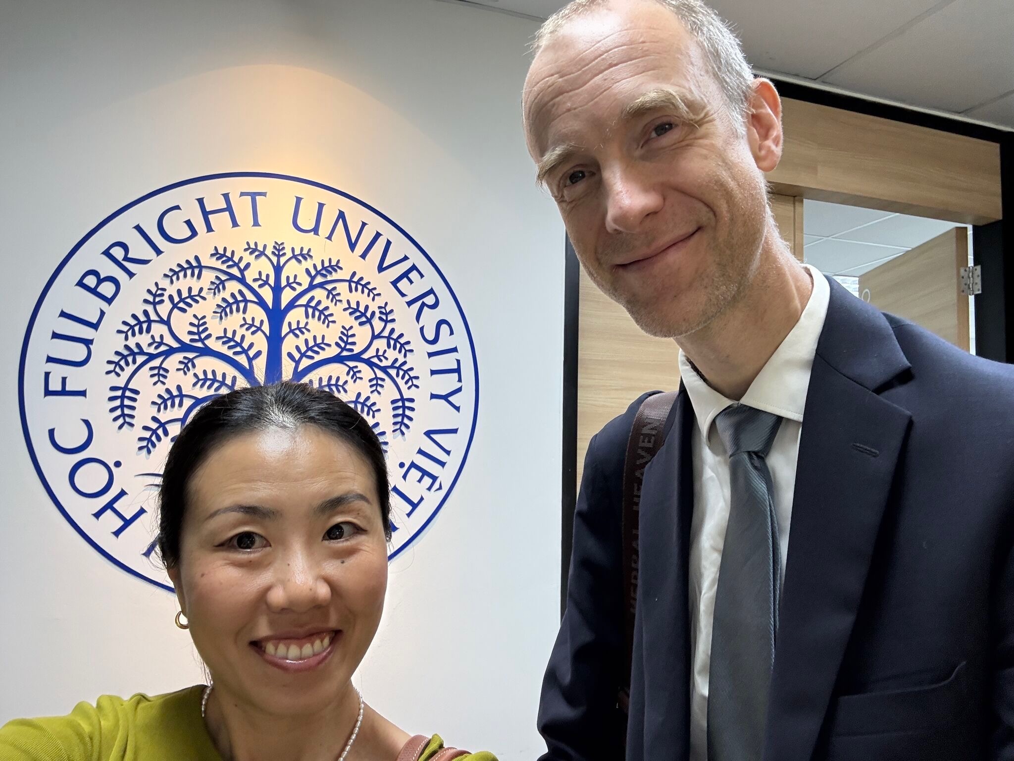 Chuck Eesley in a suit and tie standing beside a Vietnamese host in front of the Fulbright University Vietnam seal at the Crescent Campus